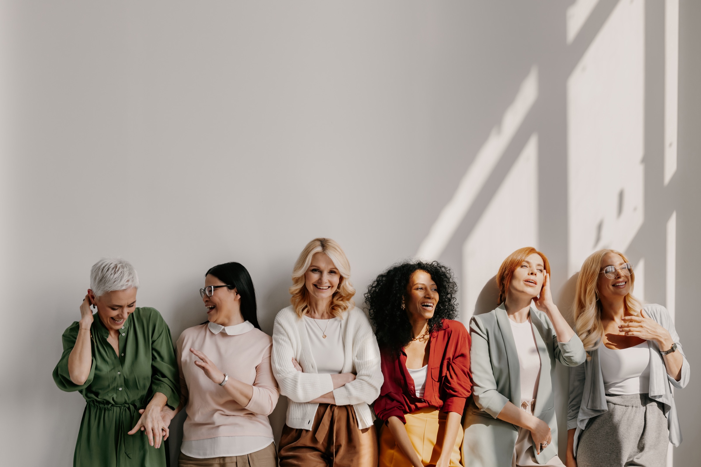 Six women of different ages laughing together against a sunlit wall